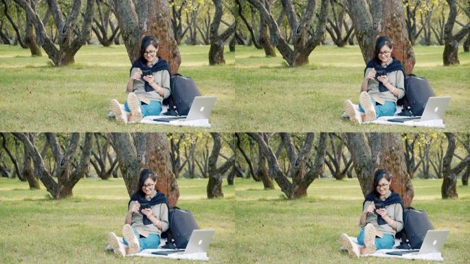 戴着眼镜微笑的年轻女子坐在公园的草地上滚动社交网络中的新闻提要