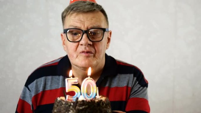 戴着蓝色眼镜的快乐成熟成年男子在生日蛋糕上吹出50支蜡烛。