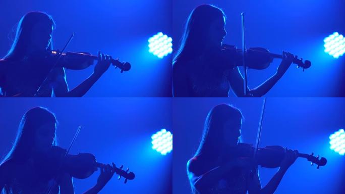一位穿着闪亮连衣裙的美丽黑发音乐家女性的剪影在一个蓝色照明的黑暗工作室里拉小提琴。特写