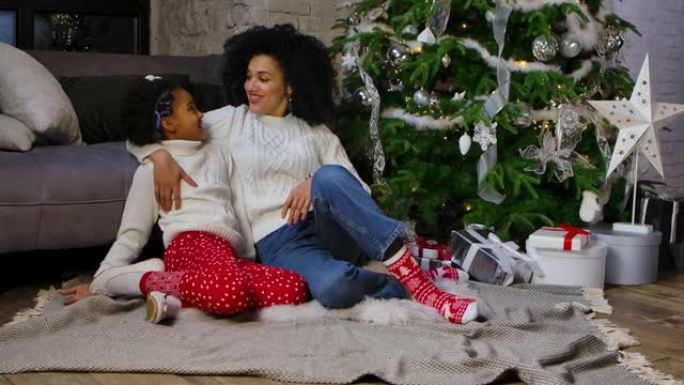 非洲裔美国妈妈和她的小女儿拥抱和说话可爱的肖像。女人和女孩坐在沙发和装饰圣诞树附近。家庭之夜快乐。慢