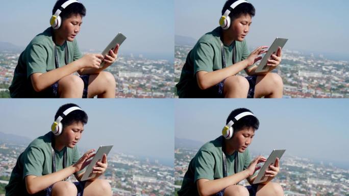 快乐的亚洲男孩戴着耳机，在阳光明媚的日子里用数字平板电脑在山顶上，有自然背景。