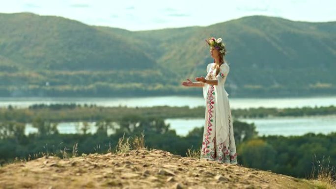 美丽的年轻女子站在小山上，头上戴着花环，穿着带有刺绣的民间服装。女人用手唱民歌和手势，大自然的广阔。