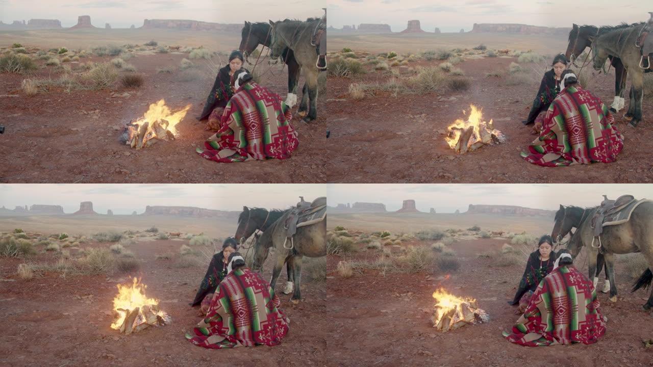 两个美洲原住民姐妹，一个在打鼓，包裹着传统的纳瓦霍人毯子，在他们身后的篝火马，标志性的纪念碑，日落