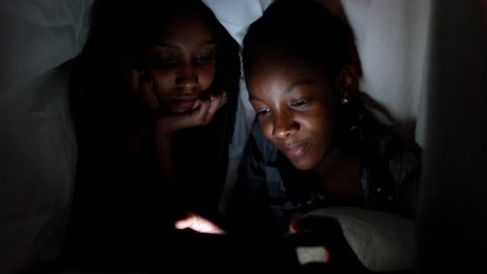 晚上在毯子下的两个姐妹在智能手机设备上观看视频