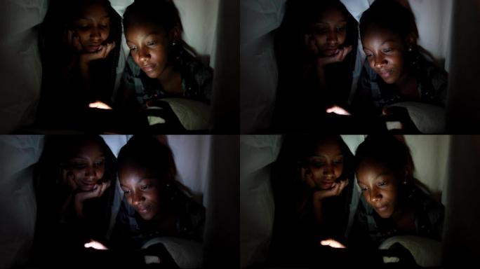 晚上在毯子下的两个姐妹在智能手机设备上观看视频