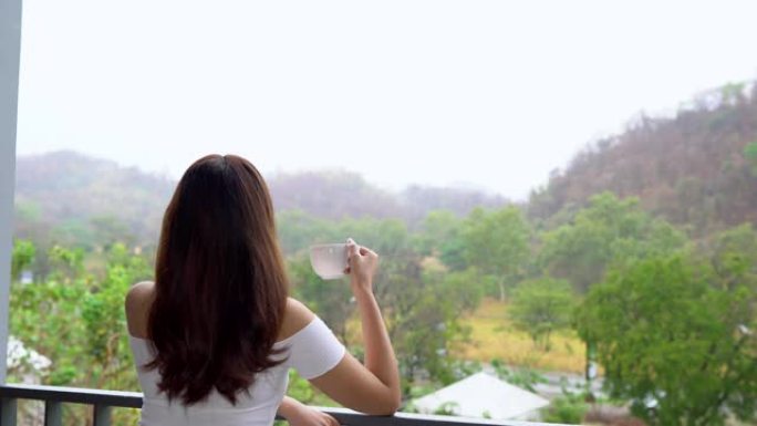 亚洲妇女喝咖啡或茶，在公寓阳台上眺望山景