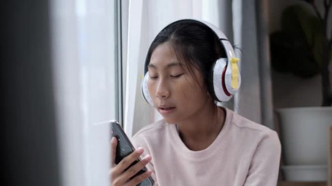 亚洲人坐在家里窗户附近的地板上时，使用智能手机和耳机唱歌。
