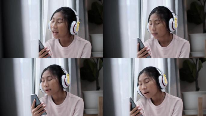 亚洲人坐在家里窗户附近的地板上时，使用智能手机和耳机唱歌。