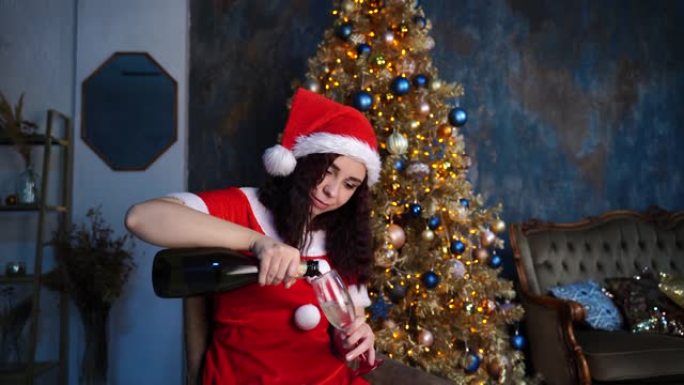 穿着圣诞老人套装的年轻女子坐在圣诞树附近的椅子上，将香槟倒入玻璃杯中。迷人的黑发女人用酒精填充透明玻