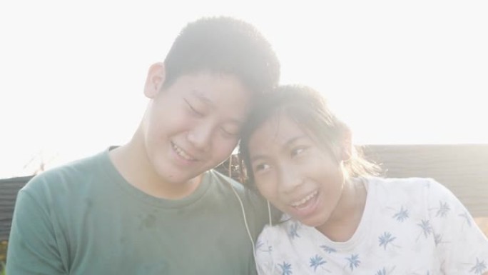 快乐的亚洲男孩为他的妹妹分享耳塞，并与太阳光背景，技术概念一起听歌。