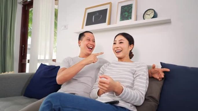 30多岁的年轻成年丈夫和妻子坐在沙发上时使用遥控器打开电视的肖像。亚洲幸福的夫妇呆在家里，在舒适的客