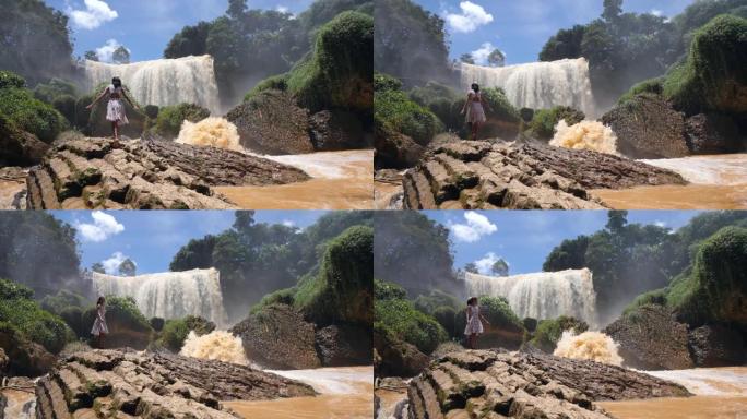 穿着碎花连衣裙的年轻女子在巨大的瀑布流附近的一块巨大岩石上行走
