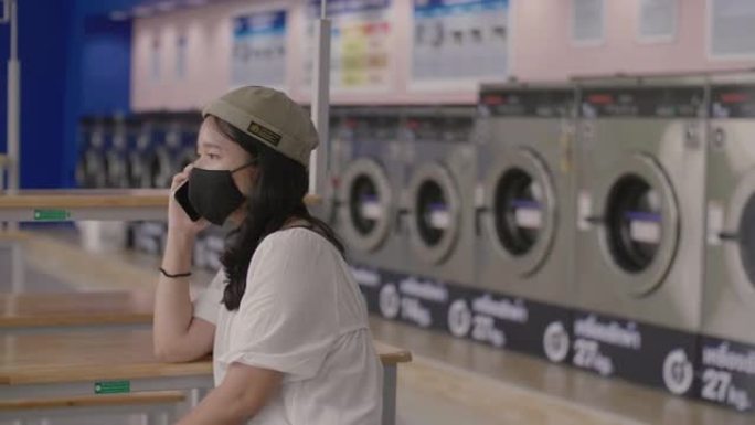 亚洲女孩手里拿着智能手机时戴着maskat。她在自动洗衣店打电话。