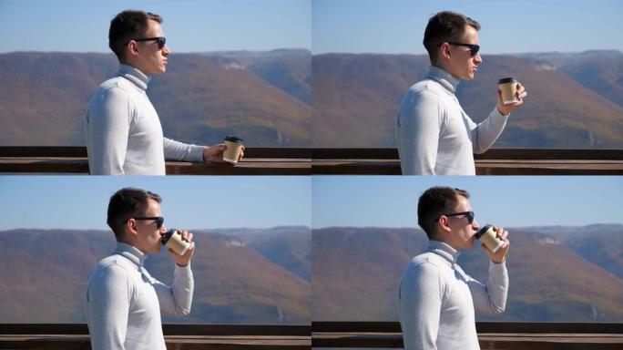 在晴朗的日子里，穿着灰色高领毛衣和太阳镜的年轻人从山底上的塑料玻璃杯中喝一杯饮料