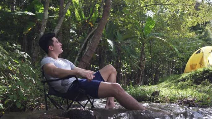4k快乐英俊的亚洲男人喜欢在森林徒步旅行和露营。健康的家伙坐在小溪边的椅子上，与美丽的大自然放松。户