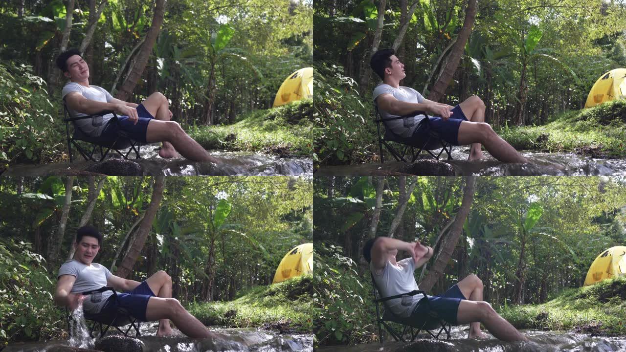 4k快乐英俊的亚洲男人喜欢在森林徒步旅行和露营。健康的家伙坐在小溪边的椅子上，与美丽的大自然放松。户