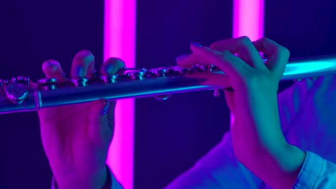 由专业长笛演奏家演奏的古典音乐交响音乐会。女性手触摸长笛的按键特写。明亮的紫色霓虹灯背景下的管乐器。