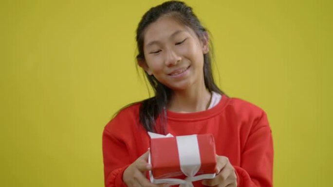 穿着红色毛衣的快乐亚洲女孩拿着黄色背景的礼品盒，庆祝节日或生日概念。