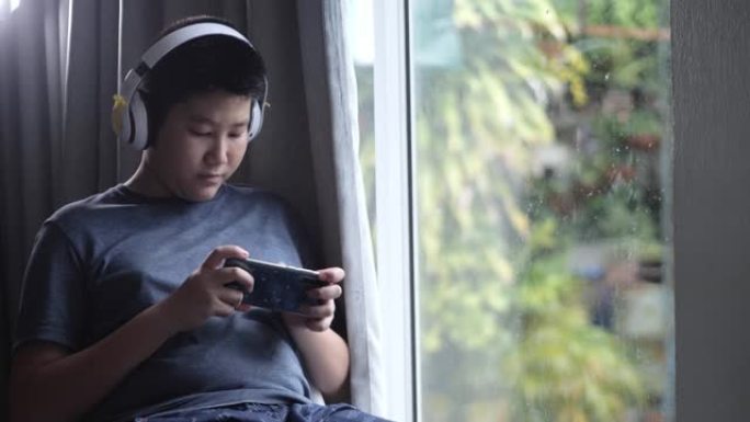 亚洲男孩坐在雨天附近的椅子上，男孩通过智能手机玩网络游戏。