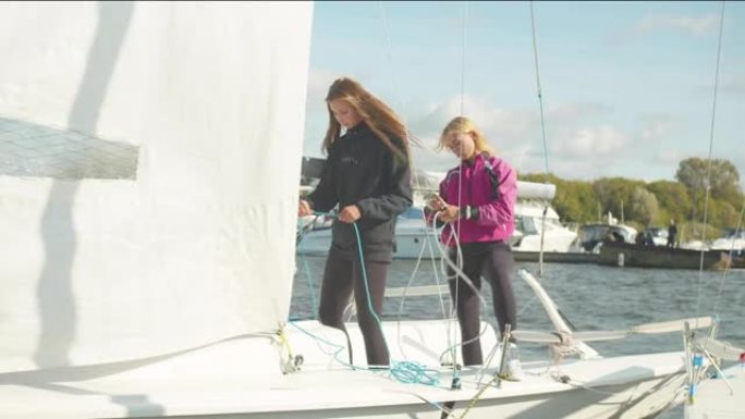 两个可爱的女孩站在一艘白色游艇的船尾，拉着绳索控制帆