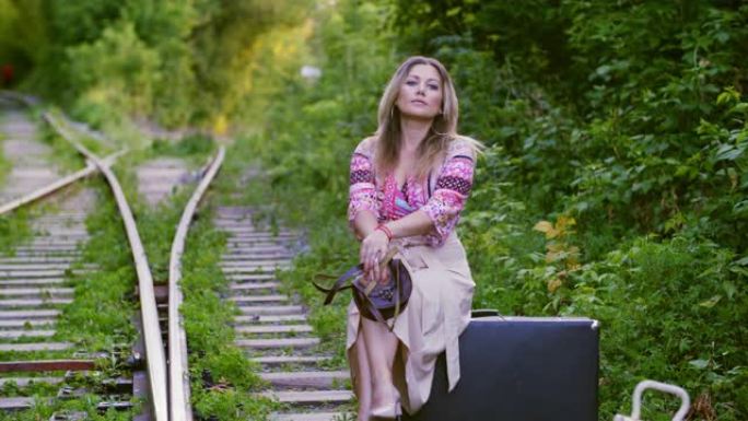 嬉皮女人坐在夏季森林铁路背景上的复古手提箱上。等待的女人坐在铁路上的老式手提箱上，看着远处。
