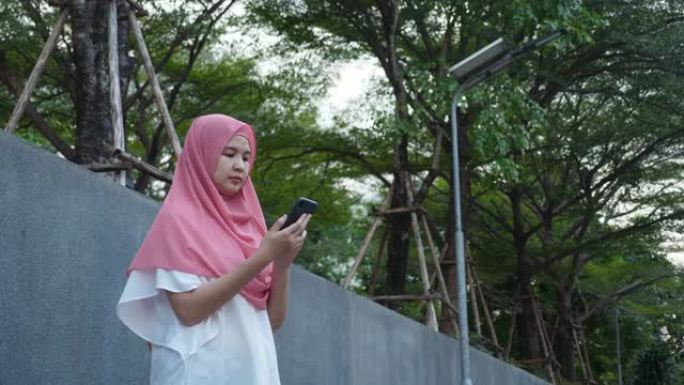 穿着粉红色头巾的漂亮的中年亚洲穆斯林妇女，在户外等出租车，用智能手机抢出租车，寻找乘车路线，挥手示意