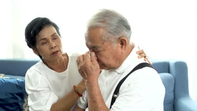 不幸的退休亚洲老年夫妇坐在沙发上，年迈的妻子在家安慰悲伤的丈夫。