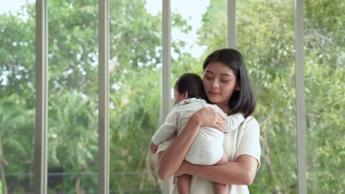 年轻的亚洲母亲站着抱着一个孩子。让他睡在房子的客厅里