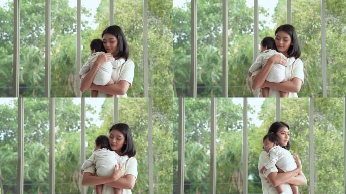 年轻的亚洲母亲站着抱着一个孩子。让他睡在房子的客厅里