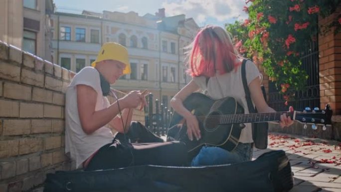 几个十几岁的潮人街头音乐家通过音乐赚钱，女孩用彩色头发唱歌和弹吉他，盖伊在智能手机上录制视频。坐在城