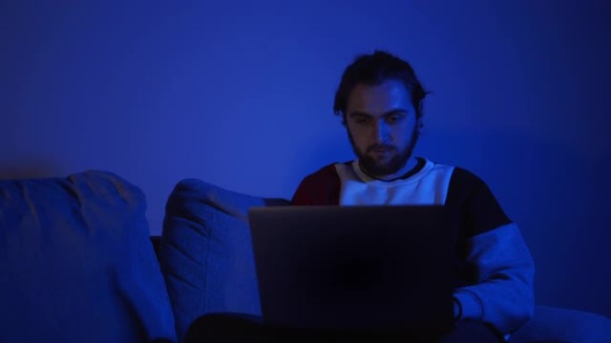昏昏欲睡和疲倦的男人在半夜坐在家里的沙发上花时间使用智能手机。年轻的男学生晚上在电脑上工作到很晚。
