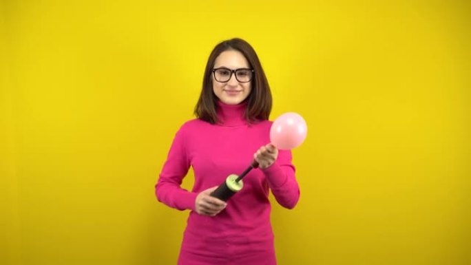 一名年轻女子用泵给一个粉红色的气球充气，它在黄色背景上爆裂。穿着粉色高领毛衣和眼镜的女孩。