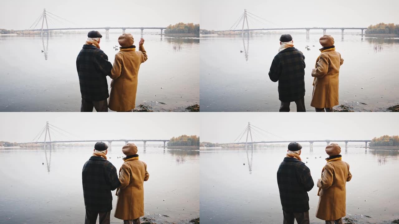 充满爱心的老年配偶在聊天，站在河岸上，喂漂浮在其中的鸭子，在秋天停车