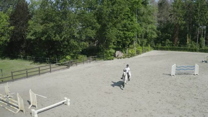 在球场上跳过障碍的白马。空中无人机射击4K。年轻的男性骑手