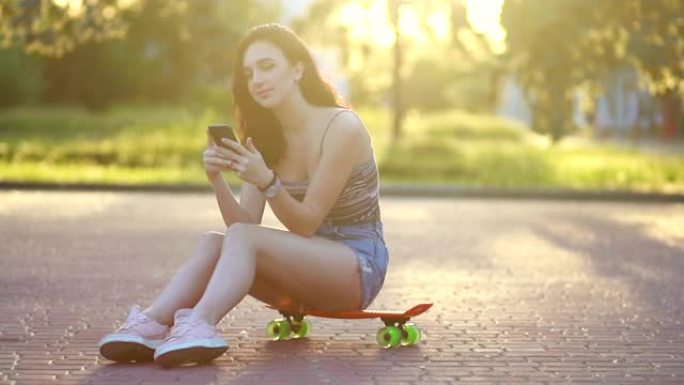 日落时一个时髦的年轻女孩微笑着坐在滑板上的肖像。