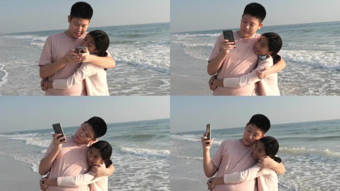 快乐的亚洲男孩和他的妹妹在假期，科技概念早上在海滩上通过智能手机与家人拍照自拍或视频通话。