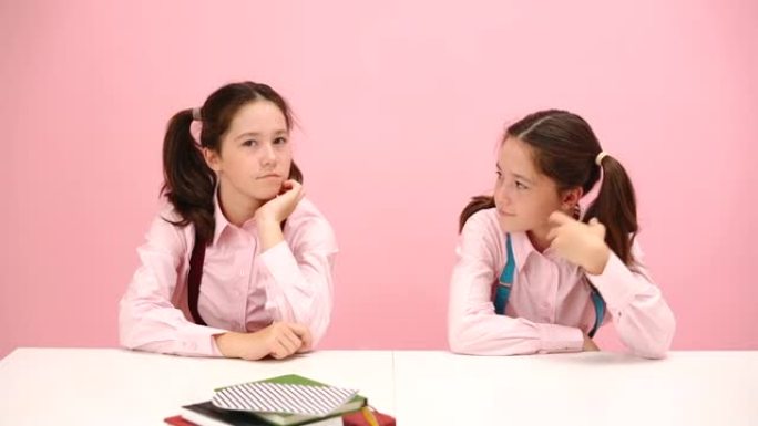快乐的双胞胎女学生坐在课桌后面。正面视图。过粉红色