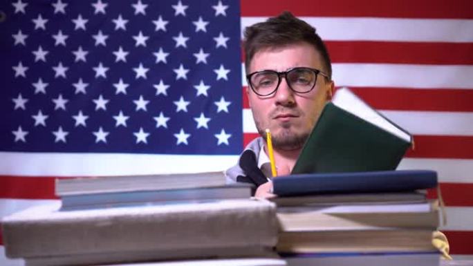 一个戴着眼镜的漂亮学生，穿着衬衫的书站在美国国旗前，在笔记本上写下想法，然后将铅笔放在一叠书上