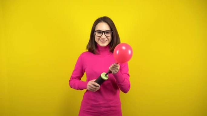 一名年轻女子用黄色背景上的泵给红色气球充气。穿着粉色高领毛衣和眼镜的女孩。
