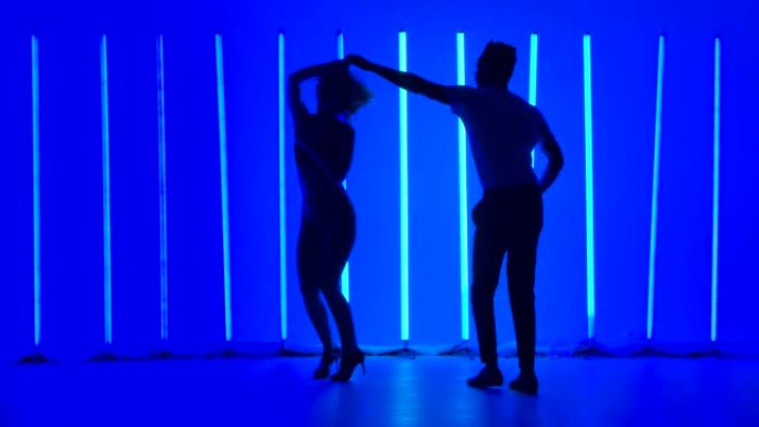 舞者在黑暗的工作室中，在明亮的蓝色霓虹灯的背景下练习一系列旋转。一个女人和一个男人在慢动作中跳舞莎莎