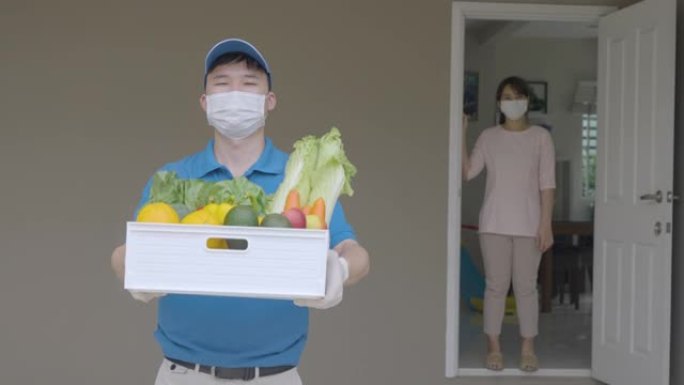 在家庭隔离期间，亚洲送货员戴着口罩和手套，上面装有食品，水果，蔬菜和饮料的食品杂物盒，站在客房门前的