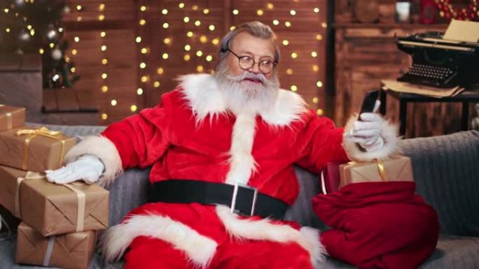 快乐的男性圣诞老人聊天视频通话在平安夜使用智能手机。4k龙红相机