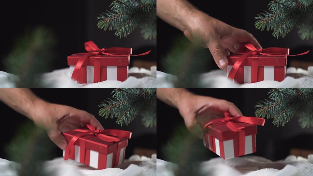 一个带有蝴蝶结的红色盒子躺在云杉树枝下的雪地里。男人的手拿礼物