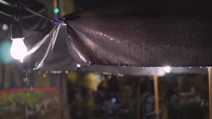 户外老城街咖啡馆晚上在倾盆大雨中遮篷。挂在街上的灯笼和灯泡。大雨倾盆而下，因为无法识别的人坐在后台的