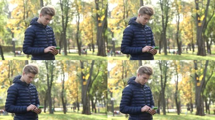 穿着蓝色夹克的年轻帅哥在秋季公园时使用手机。男人在公园的手机应用程序中阅读在线新闻并在社交网络上写短