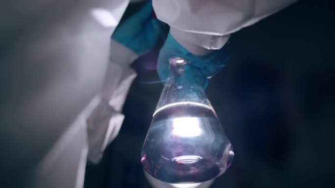 在研究实验室中进行危害实验，在对比光下用透明液体对玻璃烧杯进行特写。戴着防护手套和工作服的科学家在烧
