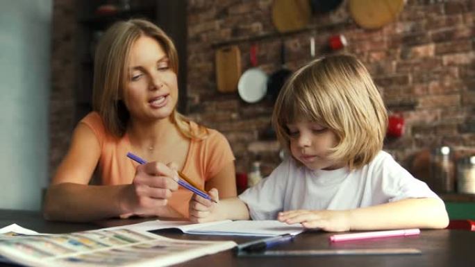 妈妈帮她的小男孩在学校字帖里做作业。她解释课程，坐在厨房里的桌子旁，室内时尚。慢动作