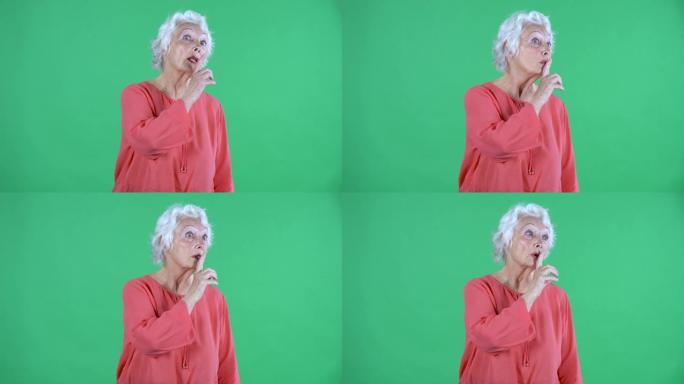 肖像老年妇女环顾四周，做一个秘密手势。工作室绿色屏幕上穿着红色上衣的白发祖母。特写