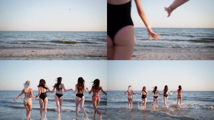 海边度假时，年轻美丽苗条的女孩们穿着泳衣，手牵着手沿着沙滩跑进水中，在夏日周末的天空和海浪的背景下
