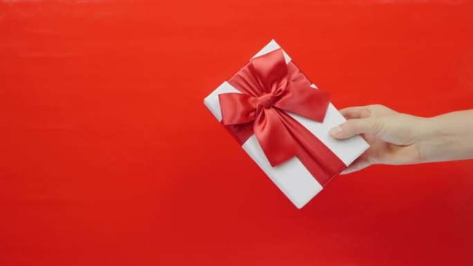 红色背景下的圣情人节礼物。情人节或生日用手展示带有红色蝴蝶结的盒子。带红丝带的白色礼品盒特写